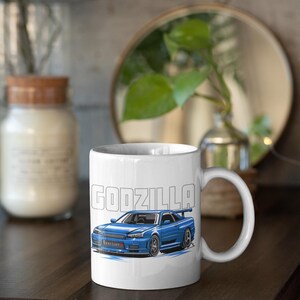 May include: White ceramic mug featuring a blue sports car illustration with the word "GODZILLA" in a bold, blocky font. The car design includes a spoiler and is set against a light blue background. The mug has a curved handle.