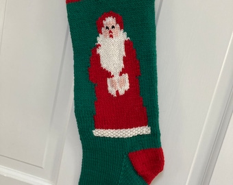 Könnte beinhalten: Ein grüner, gestrickter Weihnachtssocken mit einem roten und weißen Weihnachtsmann-Motiv und dem Wort "NOEL" in weißen Buchstaben am Bündchen.