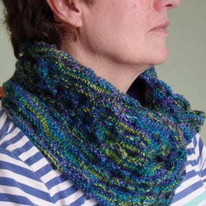 May include: A woman wearing a blue, green, and purple knitted cowl neck scarf. The scarf is made of a soft, textured yarn and has a unique, abstract pattern.