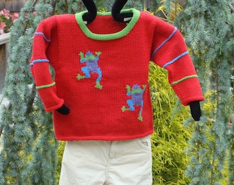 Könnte beinhalten: Roter Strickpullover mit grünen und blauen Froschmotiven. Der Pullover hängt an einem Kleiderbügel und hat lange Ärmel. Eine weiße Hose wird unter dem Pullover getragen.