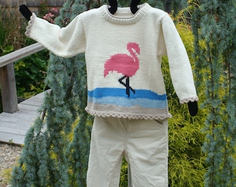 Könnte beinhalten: Ein weißer Strickpullover mit einem pinkfarbenen Flamingo-Motiv und blauen und beigefarbenen Streifen. Der Pullover hat einen Häkelrand und hängt an einem schwarzen Kleiderbügel.