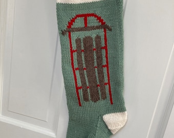 Könnte beinhalten: Ein grüner, gestrickter Weihnachtsstrumpf mit weißem Besatz und dem in Rot gestickten Wort "JOY". Der Strumpf zeigt ein braunes und rotes Schlittenmotiv.