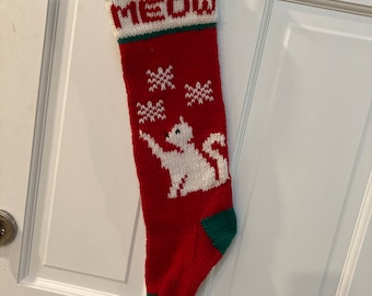 Könnte beinhalten: Rote und grüne gestrickte Weihnachtsstrumpf mit einer weißen Katze und dem Wort "MEOW" auf der Vorderseite.