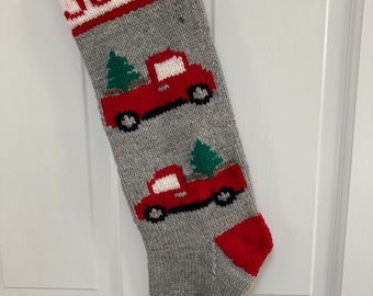 Könnte beinhalten: Ein grauer, gestrickter Weihnachtssocken mit roten und weißen Borten. Der Socken hat den Namen "JOLLY" auf der Manschette gestickt und zeigt zwei rote Lastwagen, die grüne Weihnachtsbäume transportieren.