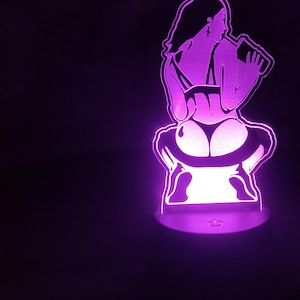 May include: A purple LED lamp featuring a silhouette of a person in lingerie drinking from a bottle. The lamp has a round base and emits a soft purple glow against a black background.