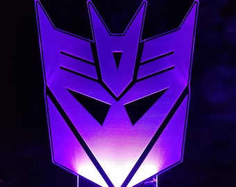 Puede incluir: Una luz nocturna iluminada de color morado y blanco con forma del logotipo de los Decepticons de la franquicia Transformers.