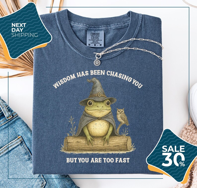 a blue t-shirt with a whimsical illustration of a frog wearing a wizard's hat and holding a book, accompanied by the text "WISDOM HAS BEEN CHASING YOU BUT YOU ARE TOO FAST". The t-shirt is displayed on a white background, with a pair of jeans and a sweater visible in the background. There are also two sale tags visible, one for the t-shirt and one for the jeans.