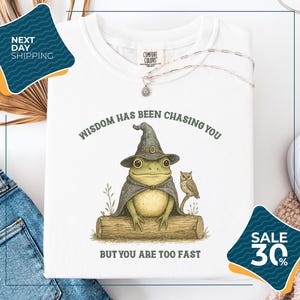 a white t-shirt with a whimsical illustration of a frog wearing a wizard's hat and holding an owl, accompanied by the text "WISDOM HAS BEEN CHASING YOU BUT YOU ARE TOO FAST". The t-shirt is displayed on a white background, with a pair of jeans and a pair of sunglasses visible in the background. There are also two sale signs in the image, one on the left side and one on the right side, indicating a discount or sale event.