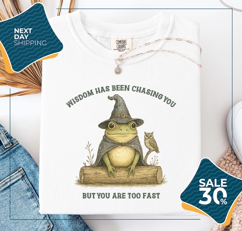 a white t-shirt with a whimsical illustration of a frog wearing a wizard's hat and holding an owl, accompanied by the text "WISDOM HAS BEEN CHASING YOU BUT YOU ARE TOO FAST". The t-shirt is displayed on a white background, with a pair of jeans and a pair of sunglasses visible in the background. There are also two sale signs in the image, one on the left side and one on the right side, indicating a discount or sale event.