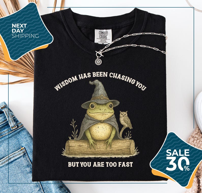 a black t-shirt with a whimsical graphic of a frog wearing a wizard's hat and holding a book, accompanied by the text "WISDOM HAS BEEN CHASING YOU BUT YOU ARE TOO FAST". The t-shirt is displayed on a white background, with a pair of jeans and a necklace visible in the foreground. There are also two sale tags in the image, one for 30% off and another for next day shipping.