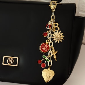 K&ouml;nnte beinhalten: Schwarze Handtasche mit goldenem Anh&auml;nger. Der Anh&auml;nger zeigt eine Sonne, Kirschen, ein herzf&ouml;rmiges Medaillon und die Worte "Be Free" auf rotem Grund. Die Tasche hat ein goldenes Logo.