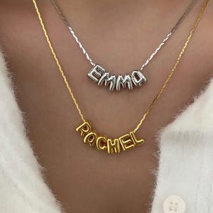Könnte beinhalten: Zwei Halsketten mit personalisierten Namensanhängern. Die obere Halskette ist silberfarben und buchstabiert "EMMA". Die untere Halskette ist goldfarben und buchstabiert "RACHEL". Beide Halsketten werden auf einem weißen Pullover getragen.