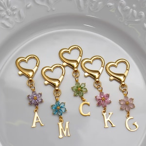 May include: Gold-coloured heart-shaped keychains with floral accents and initial charms. Each keychain features a different coloured flower and a letter charm. The keychains are displayed on a white plate.