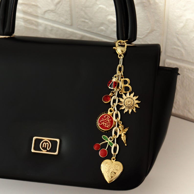 K&ouml;nnte beinhalten: Schwarze Handtasche mit goldenem Anh&auml;nger. Der Anh&auml;nger zeigt eine Sonne, Kirschen, ein herzf&ouml;rmiges Medaillon und die Worte "Be Free" auf rotem Grund. Die Tasche hat ein goldenes Logo.