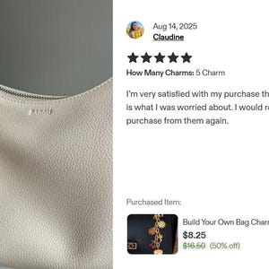 K&ouml;nnte beinhalten: Eine cremefarbene Handtasche mit einem dekorativen Anh&auml;nger. Der Anh&auml;nger hat eine goldene Kette mit verschiedenen Charms, darunter ein roter rechteckiger Charm, eine Kirsche und ein herzf&ouml;rmiger Charm. Die Tasche hat einen Rei&szlig;verschluss.