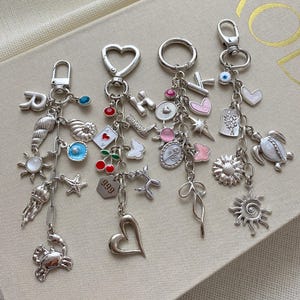 May include: Four silver keychains with various charms, including a seashell, heart, sun, and crab. Each keychain has a different clasp and a collection of decorative charms in various shapes and colors.