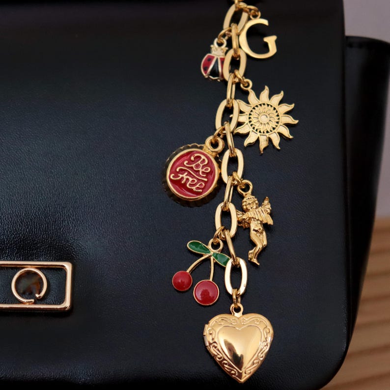 Personalized gold charm keychain and bag charm featuring a heart pendant, sun charm, cherry charm, letter initial, and decorative chain attached to a black leather bag