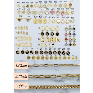 May include: A collection of gold-toned charms with various designs, including letters, numbers, zodiac signs, and other symbols. The charms are arranged in rows on a white surface. The charms are for use in jewelry making.