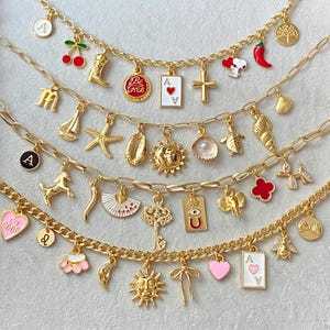 May include: Gold-toned charm necklaces with various pendants, including a cherry, a cowboy boot, a playing card, a cross, a sailboat, a starfish, a sun, a key, and a bee. The charms are suspended from a delicate chain.