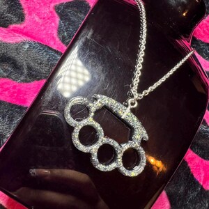 May include: A silver-toned necklace with a sparkly, silver-colored,  brass knuckle pendant. The necklace is on a black surface.