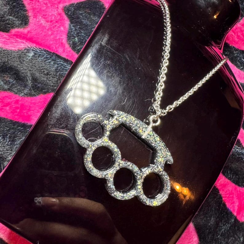 May include: A silver-toned necklace with a sparkly, silver-colored,  brass knuckle pendant. The necklace is on a black surface.