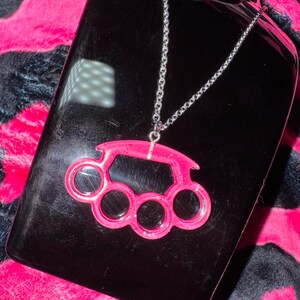 May include: A silver chain necklace with a pink plastic knuckle duster pendant. The pendant has a sparkly finish.