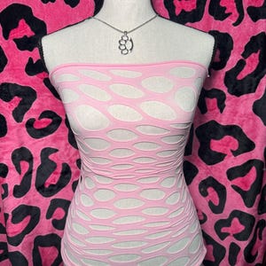 Op de afbeelding: Een roze tube top met open weefsel en een ovaal patroon. Een zilverkleurige pootafdruk hanger hangt aan een fijne ketting. De top wordt getoond op een mannequin tegen een roze luipaardprint achtergrond.