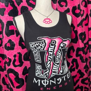 Puede incluir: Camiseta sin mangas negra con el logo de Monster Energy en rosa, blanco y negro. Un colgante de nudillos rosa cuelga de una cadena plateada. El fondo presenta un estampado de leopardo rosa y negro.