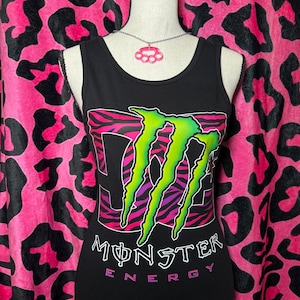 Puede incluir: Camiseta sin mangas negra con el logotipo de Monster Energy en verde neón, morado y rosa. El logotipo está sobre un fondo con estampado de cebra. Las palabras "MONSTER ENERGY" están impresas debajo del logotipo en blanco y rosa.