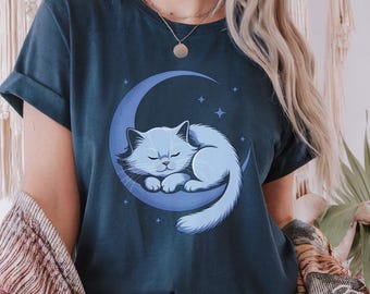 Op de afbeelding: Marineblauw T-shirt met een lichtblauwe illustratie van een slapende kat op een maan, omringd door sterren. De kat heeft gesloten ogen en een pluizige staart. Het ontwerp is gecentreerd.