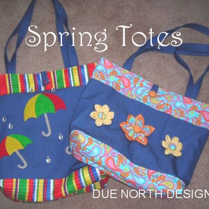 以下が含まれることがあります： カラフルな生地のアクセントが付いた青いトートバッグ2つ。1つのバッグはストライプの生地で、雨をテーマにしたデザインが施されています。もう1つのバッグはペイズリー柄で、花びらの装飾が施されています。バッグの上には青い背景に白い文字で「Spring Totes」と書かれています。バッグの下には青い背景に白い文字で「DUE NORTH DESIGNS」と書かれています。
