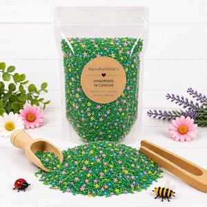 Peut inclure: Un sachet en plastique transparent rempli de garniture sensorielle verte en forme de petits bâtonnets, mélangée à de minuscules fleurs roses, jaunes et bleues. Une étiquette ronde beige indique RusticRootsPlayCo. HANDMADE IN CANADA.