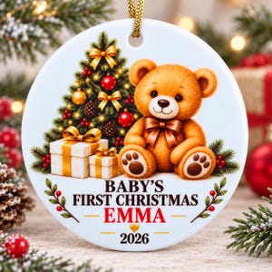 May include: A round white ceramic ornament featuring a brown teddy bear sitting next to a decorated Christmas tree and two gift boxes. The text reads BABY'S FIRST CHRISTMAS EMMA 2026, with a small gold heart icon above the year 2026.