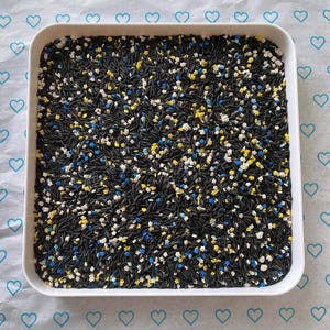 Peut inclure: Un plateau carré blanc rempli de vermicelles noirs et de petites paillettes en forme d'étoiles jaunes, blanches et bleues. Le plateau repose sur une surface à motifs bleu clair avec des cœurs.