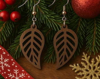 Puede incluir: Un par de pendientes de madera con forma de hoja y ganchos plateados. Los pendientes marrones tienen un diseño recortado. El fondo incluye decoraciones navideñas, como bayas rojas, una cinta roja y un copo de nieve dorado.