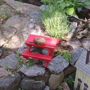 May include: A red miniature picnic table sits among rocks and greenery. A small chipmunk is perched on the table. The scene includes a miniature house and various plants, creating a whimsical garden setting.