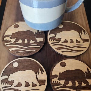 May include: Four wooden coasters featuring a bear silhouette, sun, and trees. The coasters are circular, with a light wood design and a darker wood border. A blue and white striped mug rests on a wooden surface.