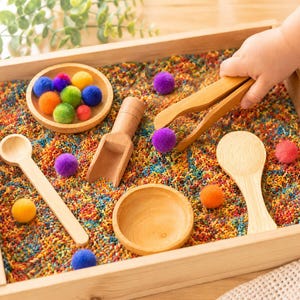 May include: A wooden sensory bin filled with multicoloured rice, featuring two small wooden bowls, a wooden scoop, a wooden spoon, and a wooden tong. Several small, fuzzy, multicoloured pom-poms are scattered throughout the bin and inside the bowls.
