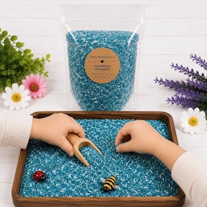 Peut inclure: Un sachet en plastique transparent rempli de riz sensoriel bleu et blanc, avec une étiquette ronde indiquant RusticRootsPlayCo. HANDMADE IN CANADA. En dessous, un plateau carré en bois contient du riz bleu, une pelle en bois, une abeille et une coccinelle.