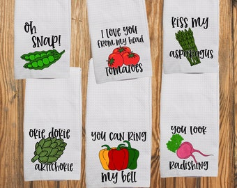 May include: Six white waffle-weave kitchen towels featuring vegetable illustrations and puns. Text includes: Oh Snap! with peas, I love you from my head tomatoes, Kiss my asparagus, Okie dokie artichokie, You can ring my bell with peppers, and You look radishing.