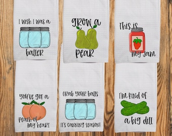 Op de afbeelding: Zes witte theedoeken met wafelstructuur, voorzien van kleurrijke illustraties en zwarte tekst. De ontwerpen bevatten weckpotten, peren, een pot aardbeienjam, perziken en augurken met humoristische teksten in zwarte letters.