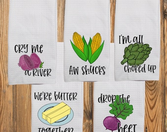 May include: Five white waffle-weave kitchen towels featuring vegetable illustrations and text. The towels display a red onion with 'CRY ME A RIVER', corn with 'AW SHUCKS', an artichoke with 'I'M ALL CHOKED UP', butter with 'WE'RE BUTTER TOGETHER', and a beet with 'DROP THE BEET'.