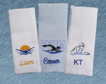 May include: Three white towels with blue and black embroidery. The towels have a swimmer graphic and the names Liam, Conner, and KT.