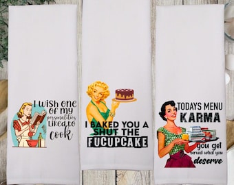 May include: Three white rectangular kitchen towels featuring retro-style illustrations and text. The first towel shows a person reading a cookbook with the text I WISH ONE OF MY PERSONALITIES LIKED TO COOK. The second shows a person holding a cake with the text I BAKED YOU A SHUT THE FUCUPCAKE. The third shows a person holding a tray with the text TODAYS MENU KARMA you get served what you deserve.