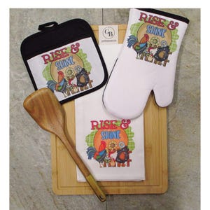 May include: A kitchen set featuring a square black-trimmed potholder, a white oven mitt, and a white towel, all printed with a rooster, sunflower, and the text "RISE & SHINE". A wooden spatula rests on a rectangular bamboo cutting board.