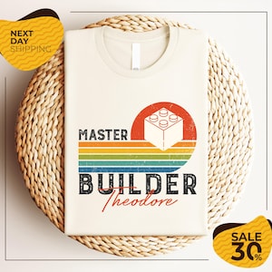 May include: A cream-colored t-shirt featuring a graphic with a white building block, a red sun, and horizontal stripes in yellow, green, and blue. The text reads MASTER BUILDER Theodore in a distressed font style, centered on the front of the shirt.