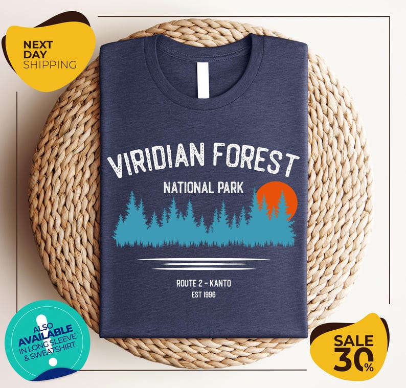 a dark blue t-shirt with a graphic design of a forest and a sunset, along with text indicating it is a t-shirt for the Viridian Forest National Park. The t-shirt is displayed on a woven basket, and there are two yellow circular stickers in the image.