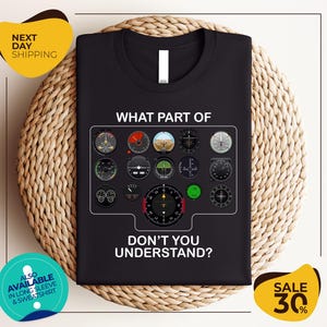 a black t-shirt with a graphic design of various flight instruments and a question asking "What part of don't you understand?". The t-shirt is displayed on a woven placemat, and there are two yellow circular banners with text around the image.