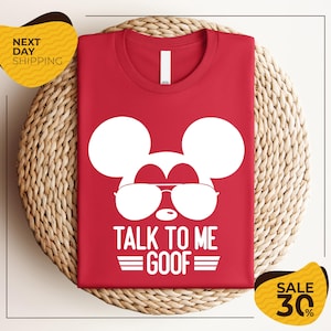 May include: Red t-shirt featuring a white graphic of Mickey Mouse wearing sunglasses, with the text "TALK TO ME GOOF". The shirt is folded on a woven surface. A yellow sticker with "NEXT DAY SHIPPING" is in the upper left corner.