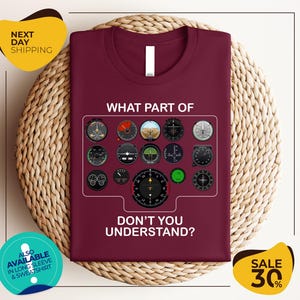 a maroon t-shirt with a graphic design of various flight instruments and a question asking "What part of don't you understand?". The t-shirt is displayed on a woven placemat, and there are two yellow circular banners with text around the image.